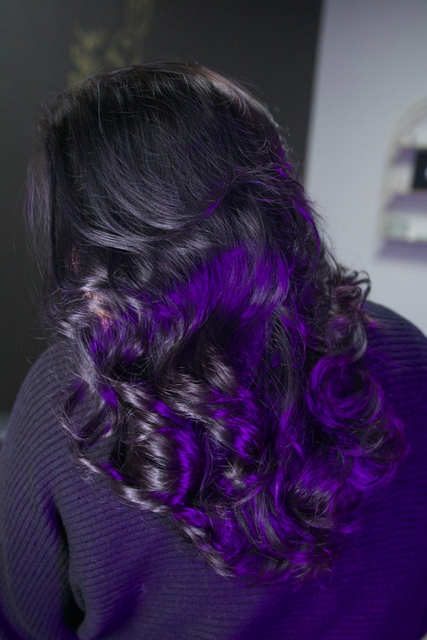 Back view of long hair with soft, long layers styled into waves under violet-tinted studio lighting.
